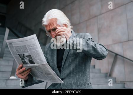 Liest Zeitung. Ein älterer Geschäftsmann in formeller Kleidung mit grauen Haaren und Bart ist draußen Stockfoto