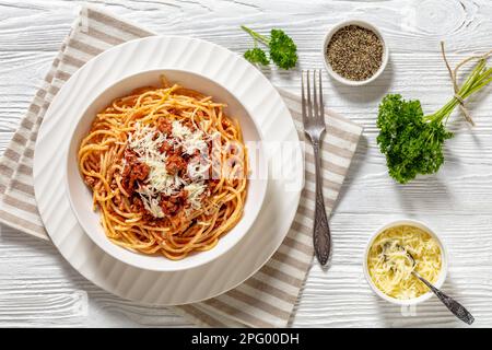 Spaghetti al Ragu alla Bolognese garniert mit geriebenem Käse in weißer Schüssel auf weißem Holztisch mit Zutaten, horizontale Sicht von oben, flach liegend Stockfoto