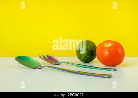 Einige schillernde Metalllöffel mit Tomaten und Avocado auf einer glatten, hellblauen Oberfläche und gelbem Hintergrund Stockfoto