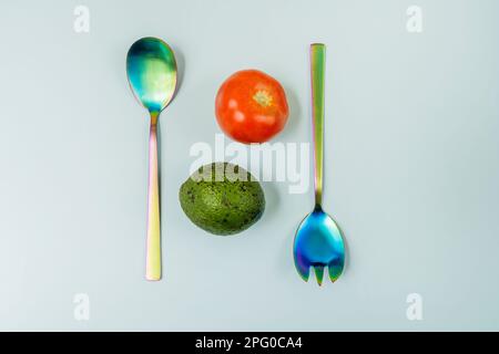 Einige schillernde Metalllöffel mit Tomaten und Avocado auf einer glatten, hellblauen Oberfläche Stockfoto