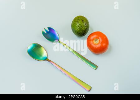 Einige schillernde Metalllöffel mit Tomaten und Avocado auf einer glatten, hellblauen Oberfläche Stockfoto