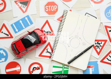 Viele verschiedene Straßenschilderkarten, Notizbuch mit Skizze des Kreisverkehrs und Spielzeugauto auf weißem Holzhintergrund, flach liegend. Fahrschule Stockfoto