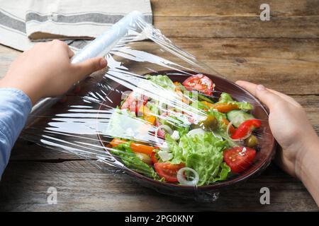 Frau, die Plastikfolie über eine Schüssel mit frischem Salat am Holztisch legt, Nahaufnahme Stockfoto