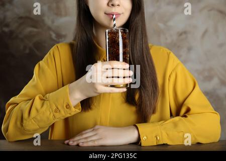 Frau trinkt Cola mit Eis auf einem Holztisch vor grauem Hintergrund, Nahaufnahme. Erfrischendes Mineralwasser Stockfoto