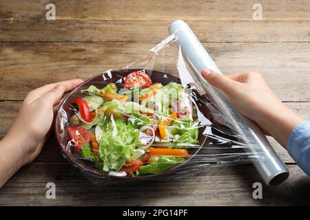 Frau, die Plastikfolie über eine Schüssel mit frischem Salat am Holztisch legt, Nahaufnahme Stockfoto