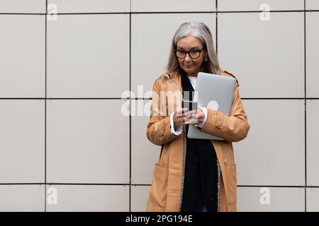 Eine Erwachsene Freiberuflerin mit grauen Haaren in Brillen, die mit einem Laptop auf der Straße stand Stockfoto