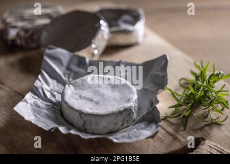 Packungen mit Camembert-Käse, von denen eines neben Rosmarin auf einem Holzbrett geöffnet ist. Stockfoto