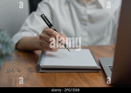 Frau Hand hält einen Stift und schreibt Informationen in ein Notizbuch, das mit einem Laptop-Computer arbeitet. Lernprogramm, Tagebuch. Stockfoto