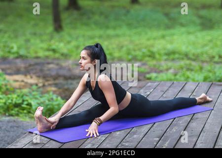 Eine junge, wunderschöne Frau in schwarzer Sportbekleidung führt die Hanumanasana-Übung durch, die Längsspalten, und trainiert allein auf einer hölzernen Terrasse im Park Stockfoto