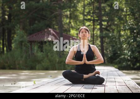 Eine Frau, die einen gesunden Lebensstil führt und Yoga praktiziert, ist in der Meditation beschäftigt, sitzt in einer Lotusposition auf einem hölzernen Pfad an einem sonnigen Sommermorgen Stockfoto