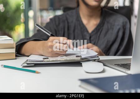 Frau Hand mit Stift, Vertrag unterschreiben, Dokument auf Klemmbrett. Büroarbeitsplatz mit Laptop und Notebook auf dem Schreibtisch. Stockfoto