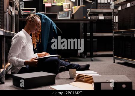 Eine afroamerikanische Polizistin, die die Akten auf der Etage durchsucht und Einblicke für die Aufklärung von Verbrechen sucht. Ermittler analysieren Hinweise und Brainstorming im Büro nachts Stockfoto