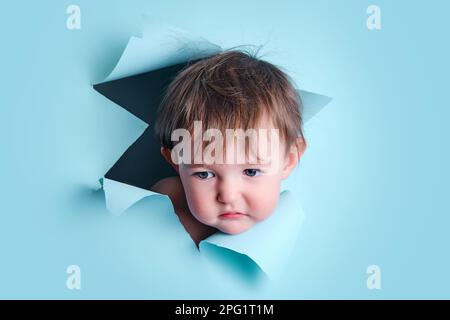 Trauriges Baby in einem Loch auf papierblauem Hintergrund. Zerrissener Studiohintergrund des Kindes, Kopierraum. Ein Kind im Alter von einem Jahr, sechs Monaten Stockfoto