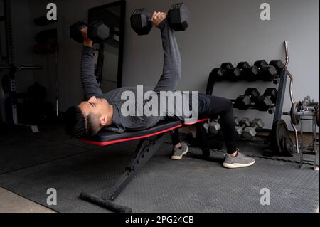 Young man Bench drückt Kurzhanteln im Fitnessstudio in der Hauswerkstatt Stockfoto