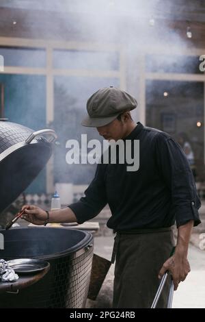 Ein asiatischer Mann in Uniform kocht draußen auf dem Grill. Küchenpersonal, Stockfoto