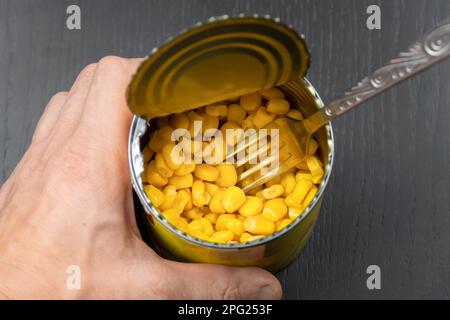 Die Hand eines Mannes hält eine offene Dose Mais und eine Gabel auf einem dunklen Tisch, Draufsicht Stockfoto