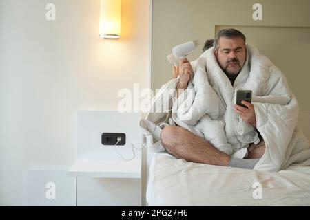 Ein Mann mittleren Alters, der auf dem Bett unter einer weißen Decke sitzt, ein Mann, der Ha benutzt Stockfoto