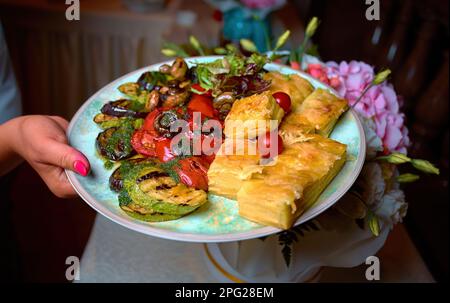 Gegrilltes Gemüse mit Pilzen und Lasagne, auf einem weißen Teller, in der Hand des Kellners Stockfoto