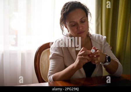 Überarbeitete, multiethnische Frau, freiberufliche Unternehmerin, die am Schreibtisch mit einem Laptop sitzt und Augentropfen in der Hand hält Stockfoto