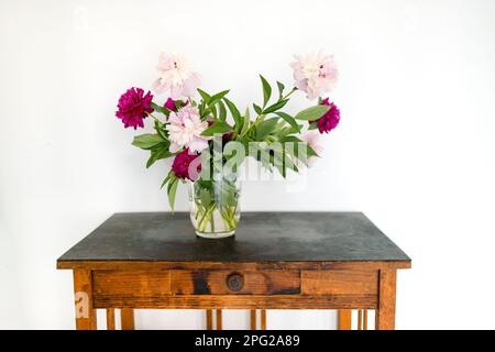 In einer transparenten Glasvase auf einem alten Holztisch stehen pinkfarbene und karmesinrote Pfingstrosen. Weißer Hintergrund. Blassende Blumen, Herbarium. Blumendesi Stockfoto