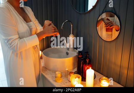 Eine junge Frau mit wunderschönem Abend-Make-up verschmiert ihre Finger mit Sahne. Hübsches Mädchen in weißen Kleidern steht im Badezimmer. Neben dem Waschbecken sind kosmetische Mittel Stockfoto