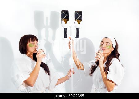 Zwei junge, brünette Frauen mit einem Glas Champagner. Zwei Flaschen Wein hängen wie medizinische Tropperinnen. Lachende weibliche Freunde in Weiß Stockfoto