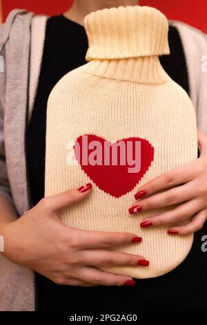 Nahaufnahme junger weiblicher Hände mit rotem Heizkissen zur Maniküre. Weißer Strickpullover mit Herz auf Heizkissen. Healthcare und valentine da Stockfoto