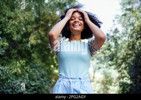 Glückliches afroamerikanisches Teenager-Mädchen in lässiger Kleidung. Lächelnde, dunkelhäutige Teenagerin, die im Park spaziert. Sommerzeit. Grüne Bäume im Hintergrund. Kopieren en Stockfoto