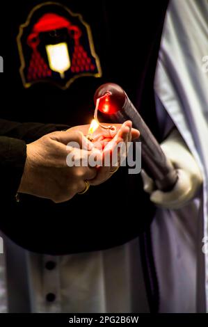 Blick auf ein Paar Hände, die eine lange rote Kerze im religiösen Stil anzünden, die während einer religiösen Veranstaltung von einer Hand mit Handschuhen gehalten wird. Stockfoto