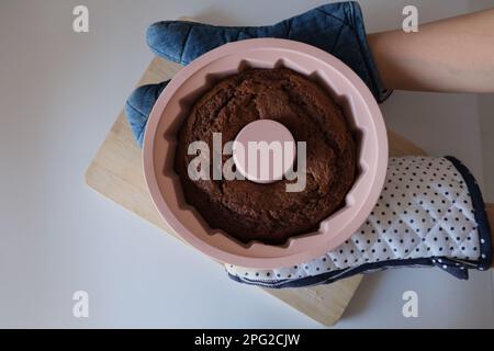 Gebackener Kuchen in einer Silikonform, Frau legt einen Kakaokuchen in einer pinkfarbenen runden Silikonform auf ein Holzbrett in der Küche. Das Konzeptfoto des Essens. Stockfoto