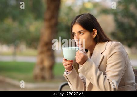 Eine Frau, die Kaffee trinkt, schaut im Winter auf den Sonnenuntergang in einem Park Stockfoto