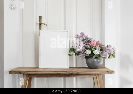 Stilleben mit Frühlingsblumen im Vintage-Stil. Leeres Bilderrahmen-Modell auf einer alten Holzbank, einem Tisch. Vase mit Flieder-, Viburnum- und Tulpenstrauß. Weiße Wand Stockfoto