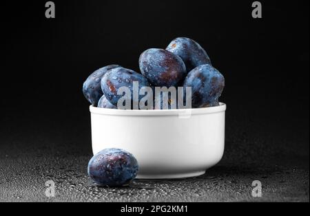 Blaue Pflaume auf einer weißen Platte mit Wassertropfen auf dunklem Hintergrund. Nützliche diätetische Früchte. Stockfoto