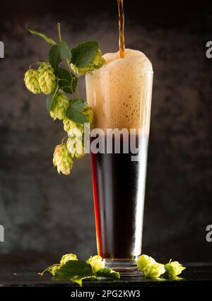 Dunkles Bier in einem Becher und grünen hop auf Holztisch auf braunem Hintergrund Stockfoto