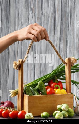 Tomaten, Gurken, Paprika, Rosenkohl, Zwiebeln und Knoblauch in einem Holzkorb. Korb mit Gemüse in den Händen auf Holzhintergrund. Stockfoto