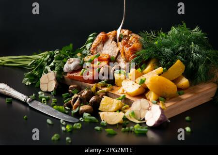 Gebratenes Hähnchen mit Kartoffeln und Pilzen auf dunklem Hintergrund. Geräuchertes Geflügel mit Gemüse und einer alten Gabel. Gebratenes Hähnchen, in Stücke geschnitten. Stockfoto