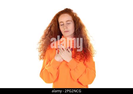 Junge Rothaarige mit orangefarbenem Pullover auf weißem Hintergrund schließt die Augen und hält die Hände auf der Brust nahe am Herzen, drückt aufrichtige Emotionen aus, Verwandtschaft zu sein Stockfoto