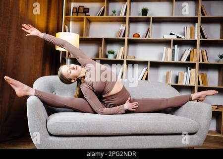 Ein süßes, sportliches Mädchen sitzt in einer gespaltenen Pose auf der Couch und streckt ihre Hand bis zum Bein. Stockfoto