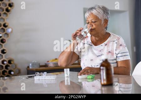 Birassische Seniorin mit grauen kurzen Haaren, die zu Hause verschriebene Medikamente und Trinkwasser einnimmt Stockfoto