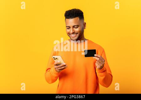 Ein sorgenfreier, glücklicher indischer Mann mit Kreditkarte und Smartphone, der auf den Smartphone-Bildschirm isoliert auf gelbem Hintergrund blickt, ein arabischer Kerl bestellt online, E-Banking-Konzept Stockfoto