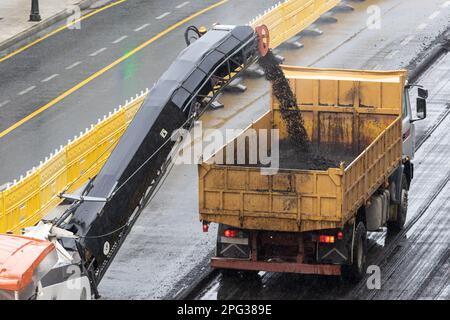 Fräsmaschine arbeitet, entfernt Asphalt von der Straße und gießt ihn in einen Lkw. Straßenbau oder Instandsetzung an regnerischen Tagen Stockfoto