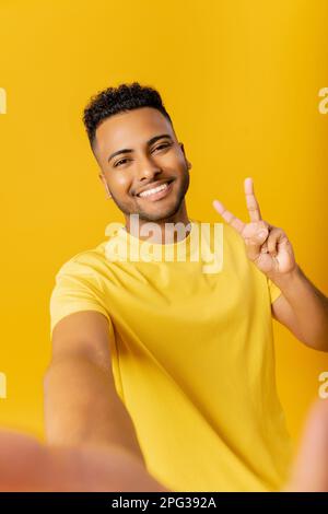 Ein witziger junger indischer Mann, der lächelt und in die Kamera schaut, Selfie auf gelbem Hintergrund macht, einen glücklichen araber, der in die Kamera lacht, eine Siegesgeste zeigt Stockfoto