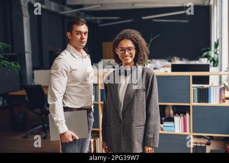Zwei junge Geschäftsleute, die im Büro zusammenstehen und vor die Kamera schauen Stockfoto