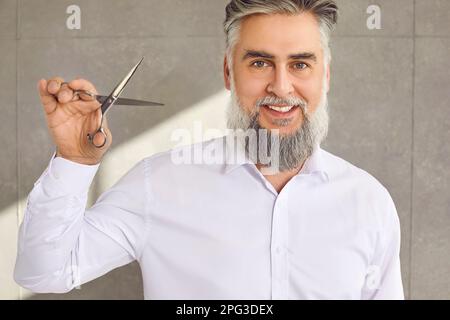 Porträt eines reifen männlichen Friseurs mit grauem Bart und Schnurrbart, der Schere hält und lächelt Stockfoto