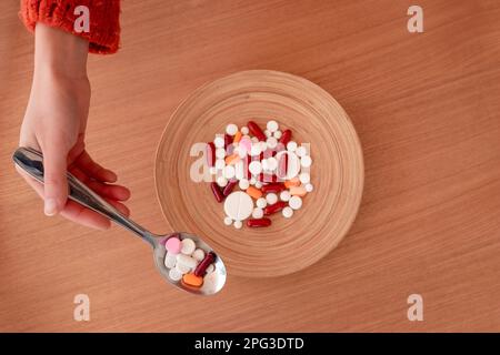 Draufsicht der anonymen weiblichen Hand, die farbige Pillen nimmt, mit Löffel vom Teller auf einem Holztisch Stockfoto