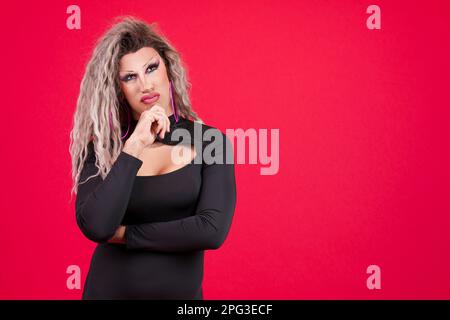 Transgender-Person mit schwarzem Kleid und Schminke mit Hand am Kinn und durchdachtem Gesichtsausdruck im Studio mit rotem Hintergrund Stockfoto