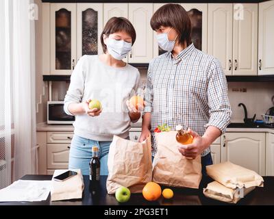 Ein junges Paar mit Schutzmasken kümmert sich um Einkäufe in der Küche. Produkte in Beuteln aus Handwerkspapier. Lebensmittellieferung. Stockfoto