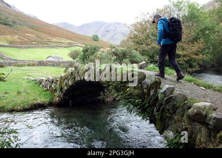 Man (Hiker) geht über die Single Stone Bogened Packhorse Bridge über Mosedale Beck in Wasdale, Lake District National Park, Cumbria, England, Großbritannien. Stockfoto