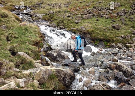 Man (Hiker) geht auf dem Gatherstone Beck Wasserfall am Gatherstone Head auf dem Black Sail Pass Path im Mosedale Valley, Lake District National Park. Stockfoto