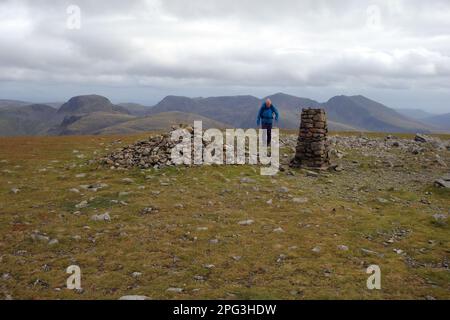 Ein Mann geht zum Steinhaufen (Cairn) & Trig Point auf dem Gipfel der Wainwright „Säule“ im Lake District National Park, Cumbria, England. Stockfoto